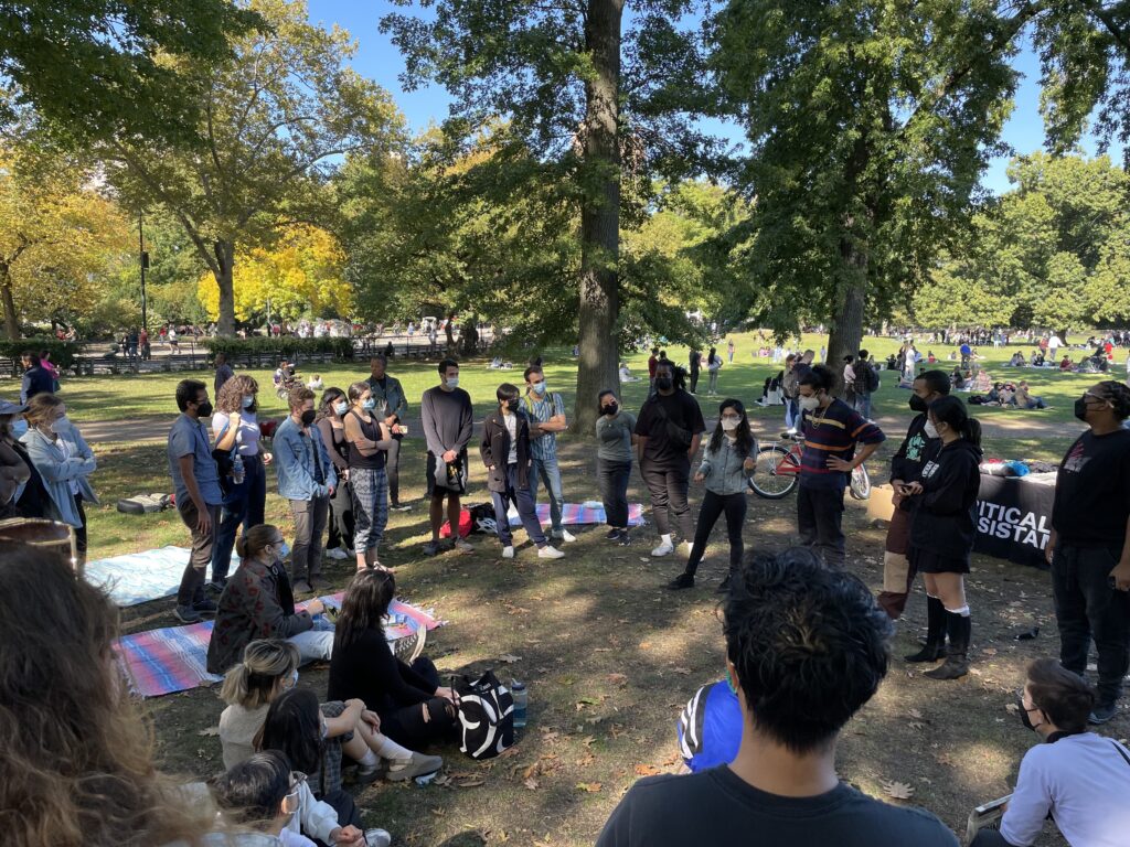 A circle of event participants standing together listening to speakers and discussing. The group is multi-gender and multi-racial. Some people are sitting down on blankets in Central Park.