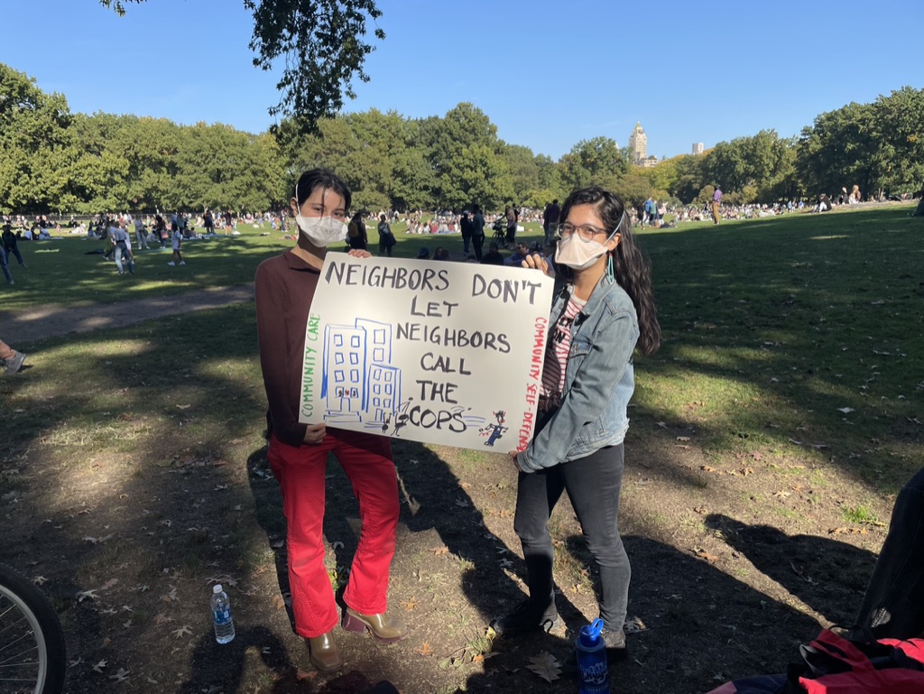 Two people with black hair holding a poster they made that says: "Neighbors Don't Let Neighbors call the cops." There's an image of a New York City apartment building next to the message. 